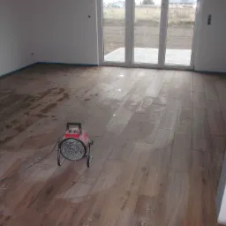 Suszenie drewnopodobnej podłogi po świeżo wykonanych pracach wykończeniowych, widoczny przenośny grzejnik elektryczny, zabezpieczone taśmą malarską ściany i duże okno balkonowe.