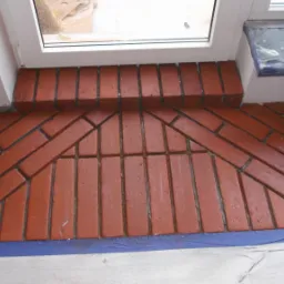 Wykończenie wnęki okiennej cegłą klinkierową ułożoną w jodełkę, zabezpieczone niebieską taśmą malarską.