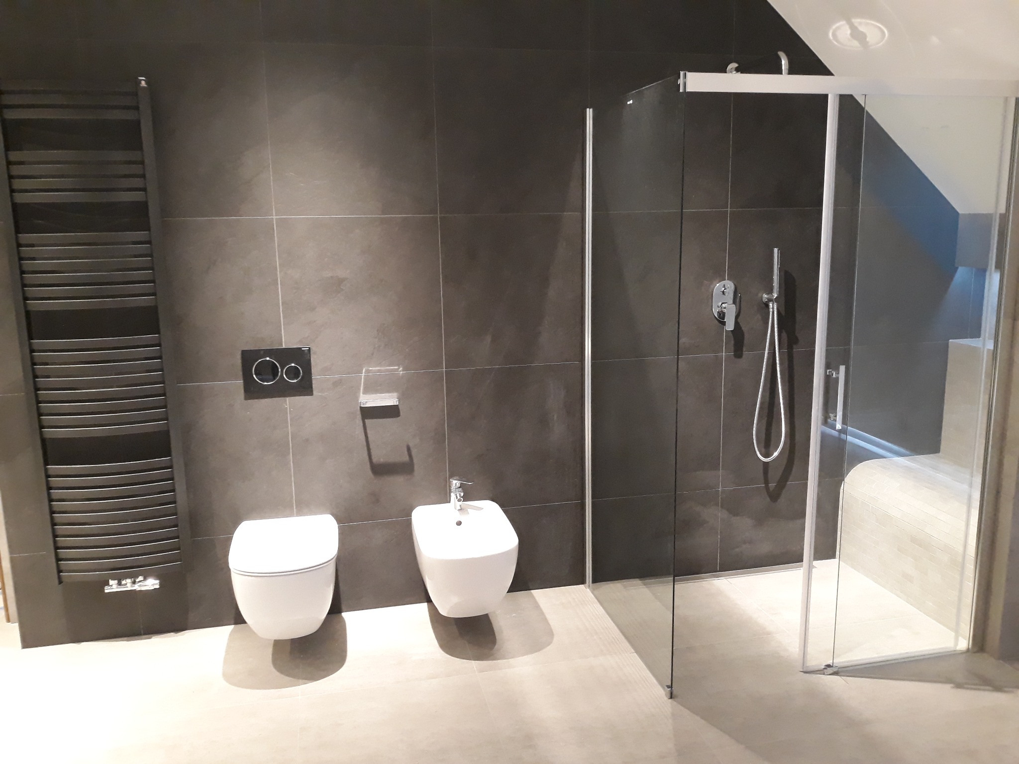 Nowoczesna łazienka w odcieniach szarości z wiszącą toaletą, bidetem i szklaną kabiną prysznicową z siedziskiem, widoczny grzejnik drabinkowy.