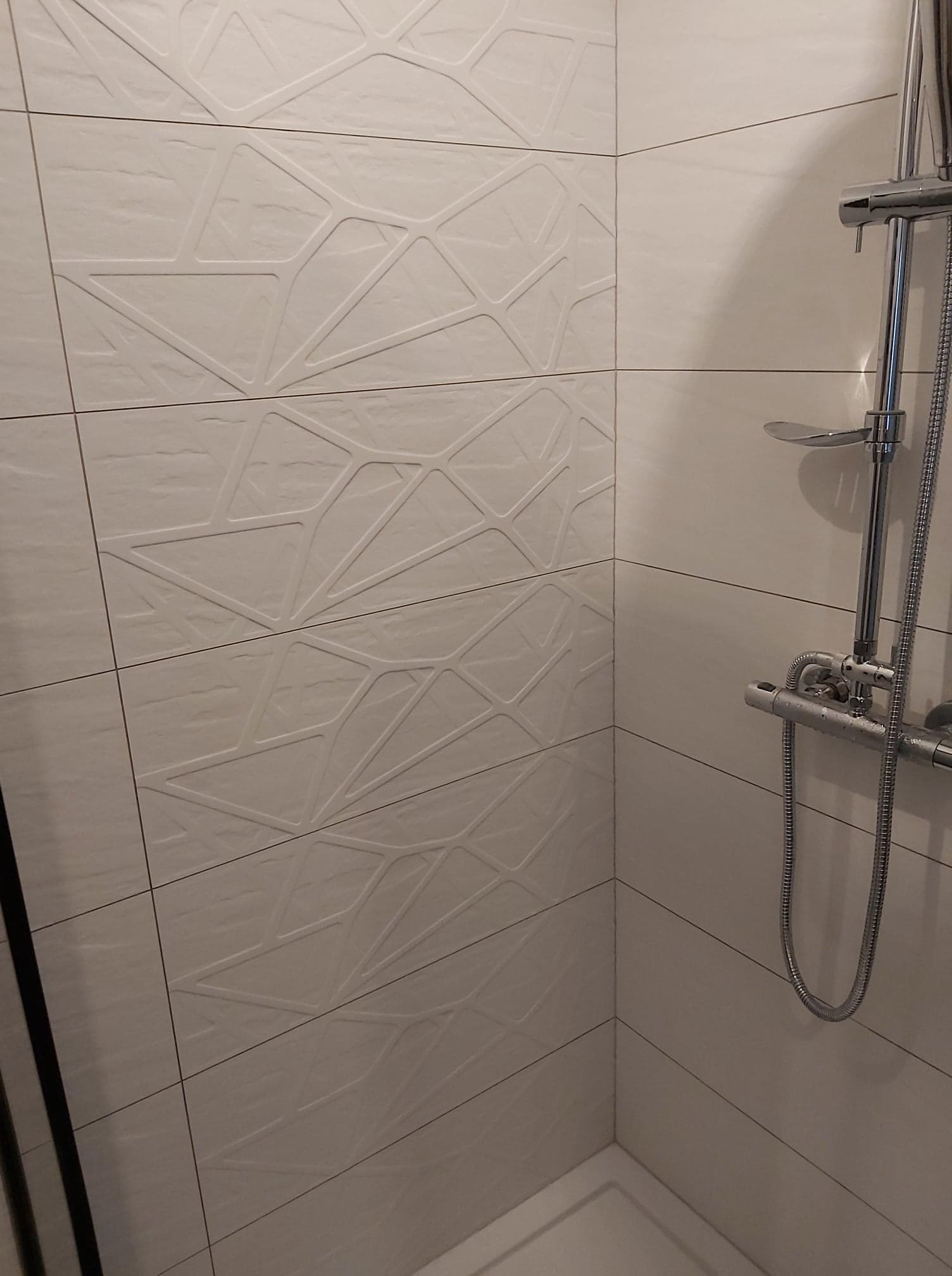 Białe, matowe płytki ścienne z geometrycznym wzorem 3D w kabinie prysznicowej z chromowaną armaturą.