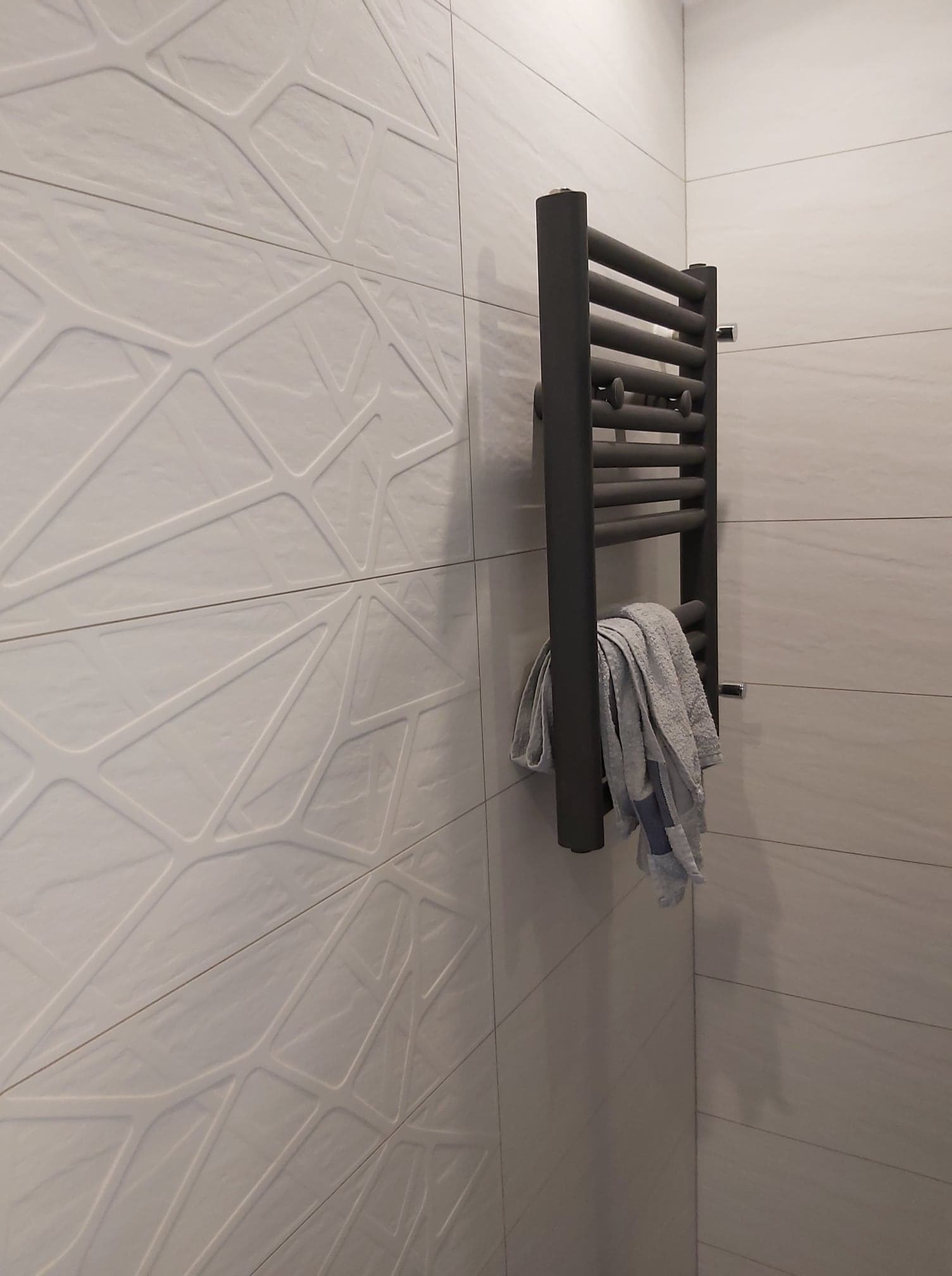 Szare, żeberkowe grzejniki łazienkowe na tle białych płytek z geometrycznym wzorem, wiszący ręcznik.