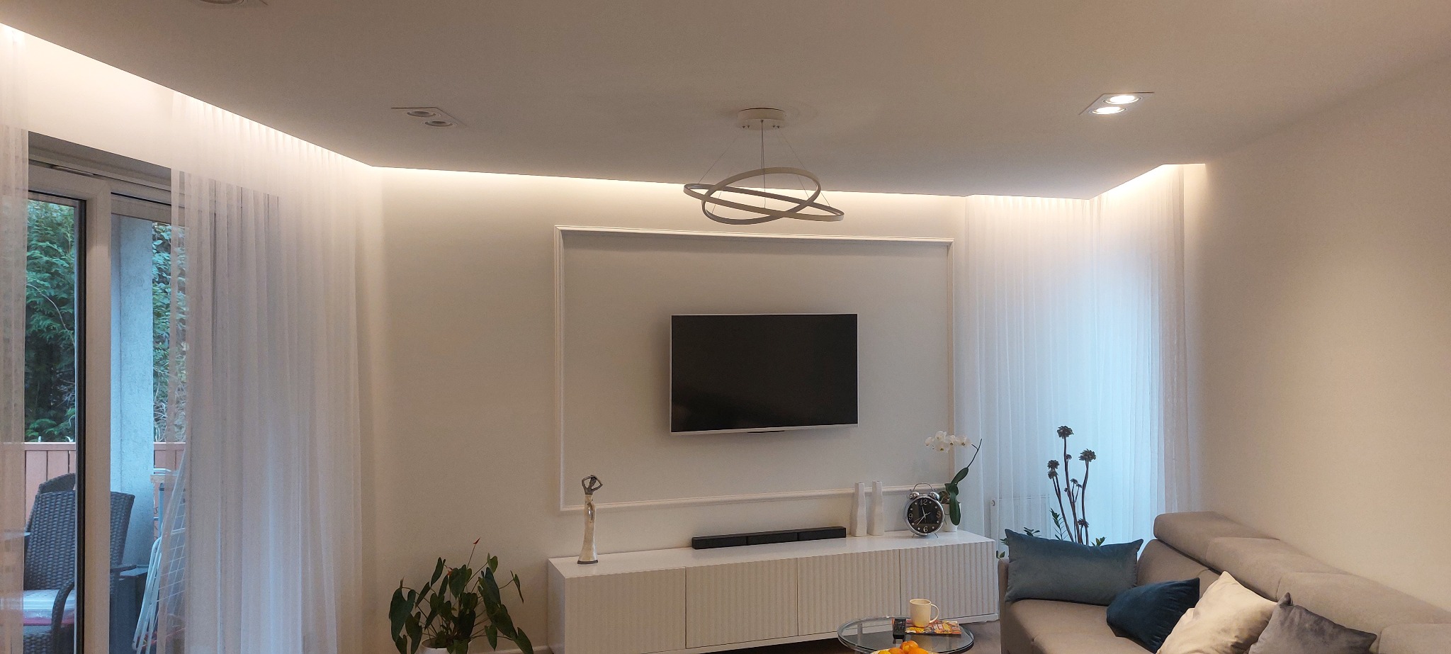 Jasne wnętrze salonu z telewizorem na ścianie, białą komodą, szarym narożnikiem i dekoracyjnym oświetleniem LED w suficie.