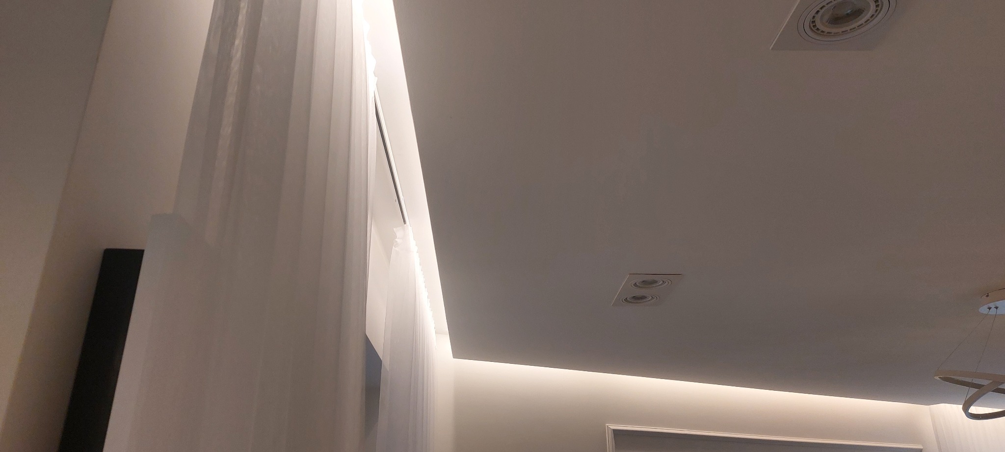 Nowoczesne oświetlenie sufitowe z wpuszczanymi lampami i ukrytym paskiem LED, podkreślającym krawędź sufitu podwieszanego; zasłony w tle.