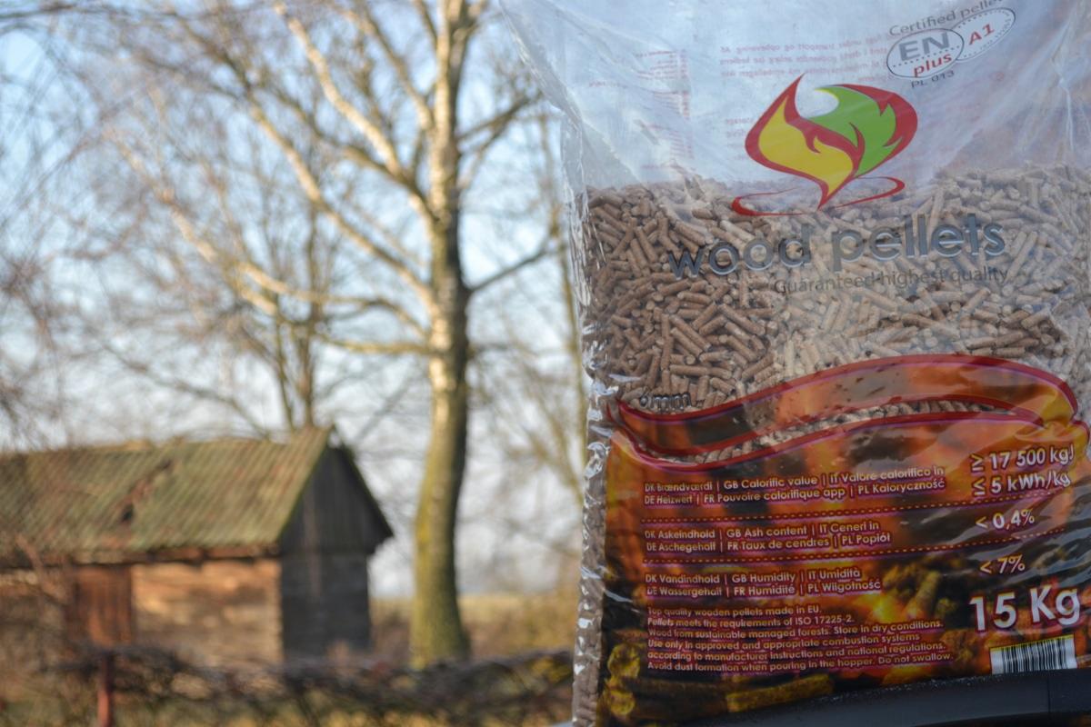 Worek 15 kg pelletu drzewnego z widocznym certyfikatem EN A1, na tle rozmytego krajobrazu z budynkiem gospodarczym i drzewami w Choroszczy.