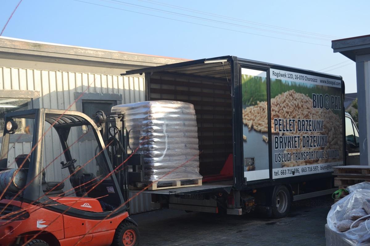 Wózek widłowy Linde ładuje paletę z towarem owiniętą folią do samochodu dostawczego z reklamą opału drzewnego na boku.