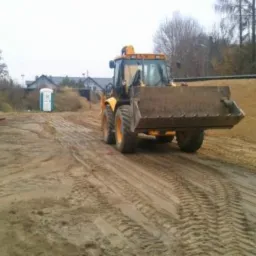 Żółta koparko-ładowarka z łyżką na placu budowy, ślady opon na piasku, w tle przenośna toaleta i zieleń. Widoczne ślady prac ziemnych.