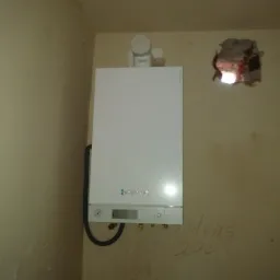 Biały piec gazowy wiszący na ścianie w surowym pomieszczeniu, widoczne ślady po instalacji i napis 'Modens 200' na ścianie.