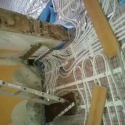 Rozbudowana instalacja ogrzewania podłogowego w trakcie montażu. Widoczne białe rurki rozprowadzone na siatce, kolektor z licznymi przyłączami oraz drabina oparta o ścianę w surowym stanie.