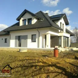 Dwukondygnacyjny dom z białym tynkiem, czarnym dachem i panelami słonecznymi, widoczny z przodu na tle błękitnego nieba z chmurami i częściowo wykończonego terenu.