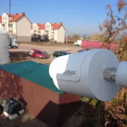 Zewnętrzna kamera monitoringu Dahua zamontowana na tle zabudowań mieszkalnych i błękitnego nieba, widoczne elementy montażowe.