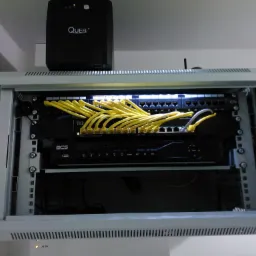 Zamknięta szafa rack z zainstalowanym switchem Cisco, rejestratorem BCS i żółtymi kablami patchcord, zasilacz awaryjny Quer na górze szafy.
