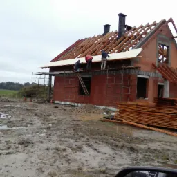 Budowa domu z czerwonej cegły w trakcie realizacji, widoczni robotnicy na rusztowaniu montujący elementy dachu, na pierwszym planie błotniste podłoże.