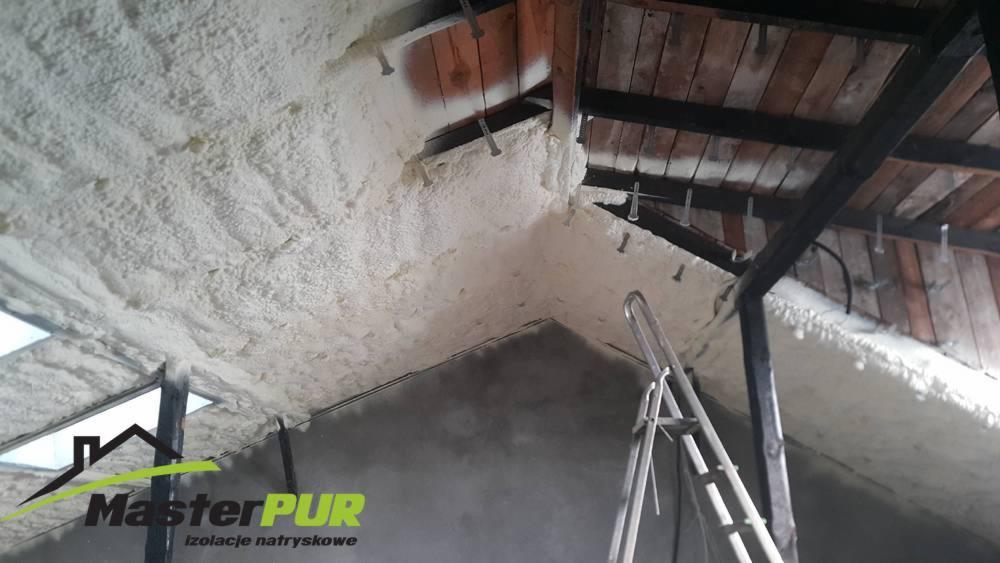 Poddasze z drewnianą konstrukcją pokryte natryskową izolacją piankową, widoczna drabina i logo firmy MasterPUR w rogu.