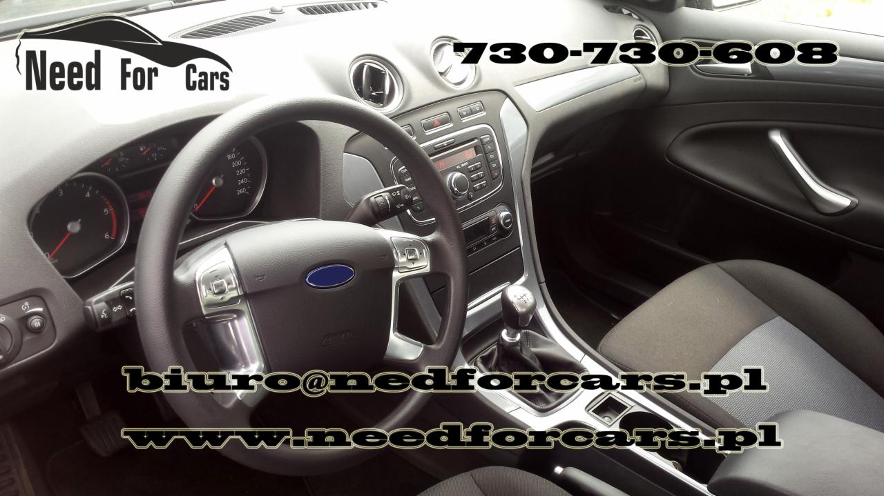 Wnętrze Forda Mondeo z widoczną deską rozdzielczą, kierownicą z logo Forda, radiem i numerem telefonu kontaktowym. Tapicerka szaro-czarna, dźwignia zmiany biegów manualna.