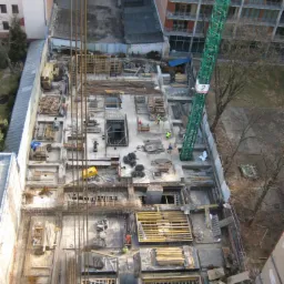 Wysoki kąt na budowę w toku: fundamenty zbrojone stalą, szalunki drewniane, dźwig budowlany. Widoczni pracownicy w kamizelkach odblaskowych.