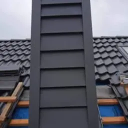 Szary komin obłożony blachą trapezową na dachu pokrytym dachówką, widoczne rusztowanie i fragment okna dachowego w pochmurny dzień.