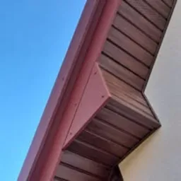 Detal dachu z brązową, drewnopodobną podbitką, widoczny fragment systemu orynnowania w kolorze bordowym oraz fragment ściany budynku na tle błękitnego nieba.
