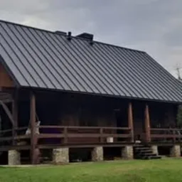 Dom z ciemnym, metalowym pokryciem dachowym, widoczne drewniane elementy konstrukcyjne i kamienne fundamenty, pochmurne niebo w tle.