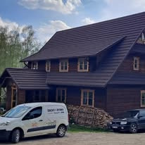 Dom z ciemnobrązowym dachem z blachy trapezowej, widoczny fragment samochodu firmowego z logo i stosem drewna obok, na tle zieleni i błękitnego nieba.