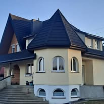 Dwupiętrowy dom z charakterystyczną wieżyczką pokrytą ciemnym dachem, widoczne nowe orynnowanie i obróbki blacharskie, jasna elewacja, schody wejściowe.