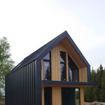 Nowoczesny dom jednorodzinny z ciemną, pionowo ułożoną blachą na elewacji i dachu, z dużymi oknami w szczycie i drewnianą okładziną na fragmentach ścian, na tle drzew.