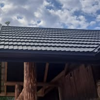 Ciemny dach z metalowej dachówki modułowej, widoczny fragment drewnianej konstrukcji budynku i błękitne niebo z chmurami w tle.