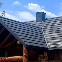 Ciemny, metalowy dach z kominem na drewnianym domu w górskim stylu, widok z dołu na tle błękitnego nieba.