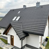 Nowoczesny dom jednorodzinny z ciemnoszarym, metalowym pokryciem dachowym imitującym dachówkę, widoczne okna dachowe i komin, biała elewacja z elementami drewnianymi.