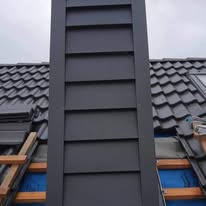 Szary komin obłożony blachą trapezową na dachu pokrytym dachówką, widoczne rusztowanie i fragment okna dachowego w pochmurny dzień.