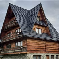 Dwupiętrowy dom z bali z ciemnoszarym dachem dwuspadowym, drewnianymi balkonami i kamienną podmurówką na tle pochmurnego nieba.