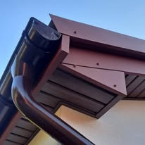 Brązowy system odprowadzania wody deszczowej, widok z dołu na narożnik dachu, z widocznymi elementami mocującymi i fragmentem elewacji budynku.