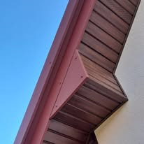Detal dachu z brązową, drewnopodobną podbitką, widoczny fragment systemu orynnowania w kolorze bordowym oraz fragment ściany budynku na tle błękitnego nieba.