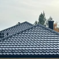 Szary dach z dachówki ceramicznej na tle pochmurnego nieba i drzew, widoczne kominy.