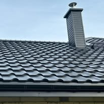 Szary dach z metalowej dachówki z kominem i rynną, widok z dołu na tle błękitnego nieba.