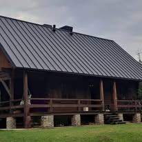 Dom z ciemnym, metalowym pokryciem dachowym, widoczne drewniane elementy konstrukcyjne i kamienne fundamenty, pochmurne niebo w tle.