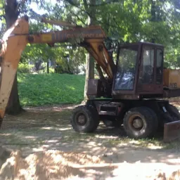 Żółto-brązowa koparka kołowa, marki Case Poclain, zagłębia łyżkę w ziemi na tle zieleni drzew i trawy w słoneczny dzień. Widoczny wykop i pryzma ziemi.
