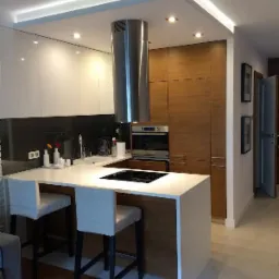 Nowoczesna kuchnia z wyspą: białe fronty szafek, drewniane wykończenia, stalowy okap. Dwa białe hokery przy wyspie. Oświetlenie LED pod sufitem.