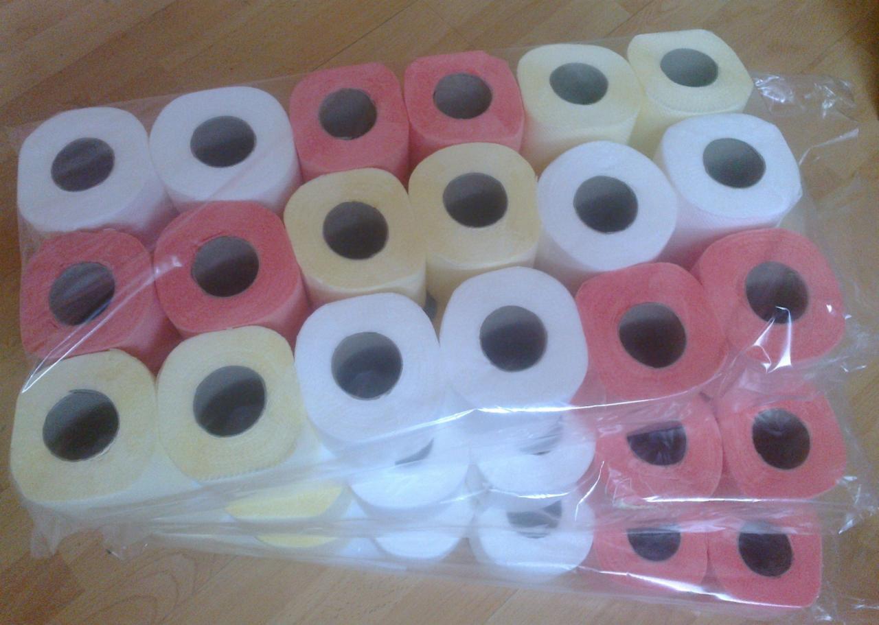 Opakowanie zbiorcze rolek papieru toaletowego w kolorach białym, żółtym i różowym, zapakowane w przezroczystą folię.