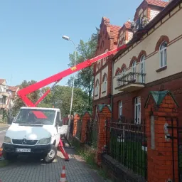 Czerwony podnośnik koszowy na białym samochodzie Mercedes-Benz Sprinter ustawiony na chodniku z pomarańczowymi pachołkami, sięgający do dachu dwupiętrowego budynku z czerwonej cegły i jasnej...