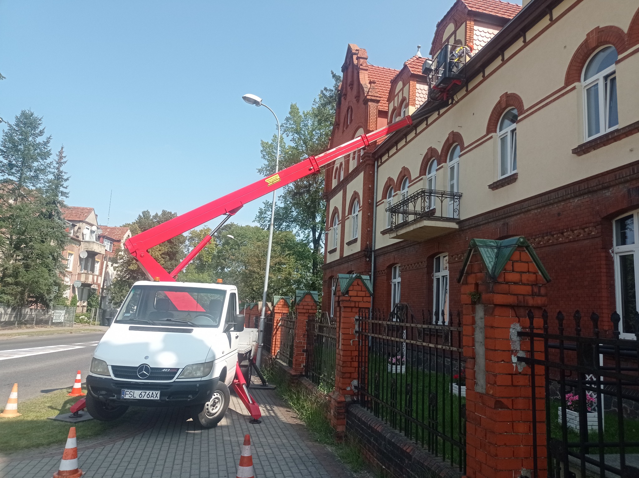 Czerwony podnośnik koszowy na białym samochodzie Mercedes-Benz Sprinter ustawiony na chodniku z pomarańczowymi pachołkami, sięgający do dachu dwupiętrowego budynku z czerwonej cegły i jasnej...