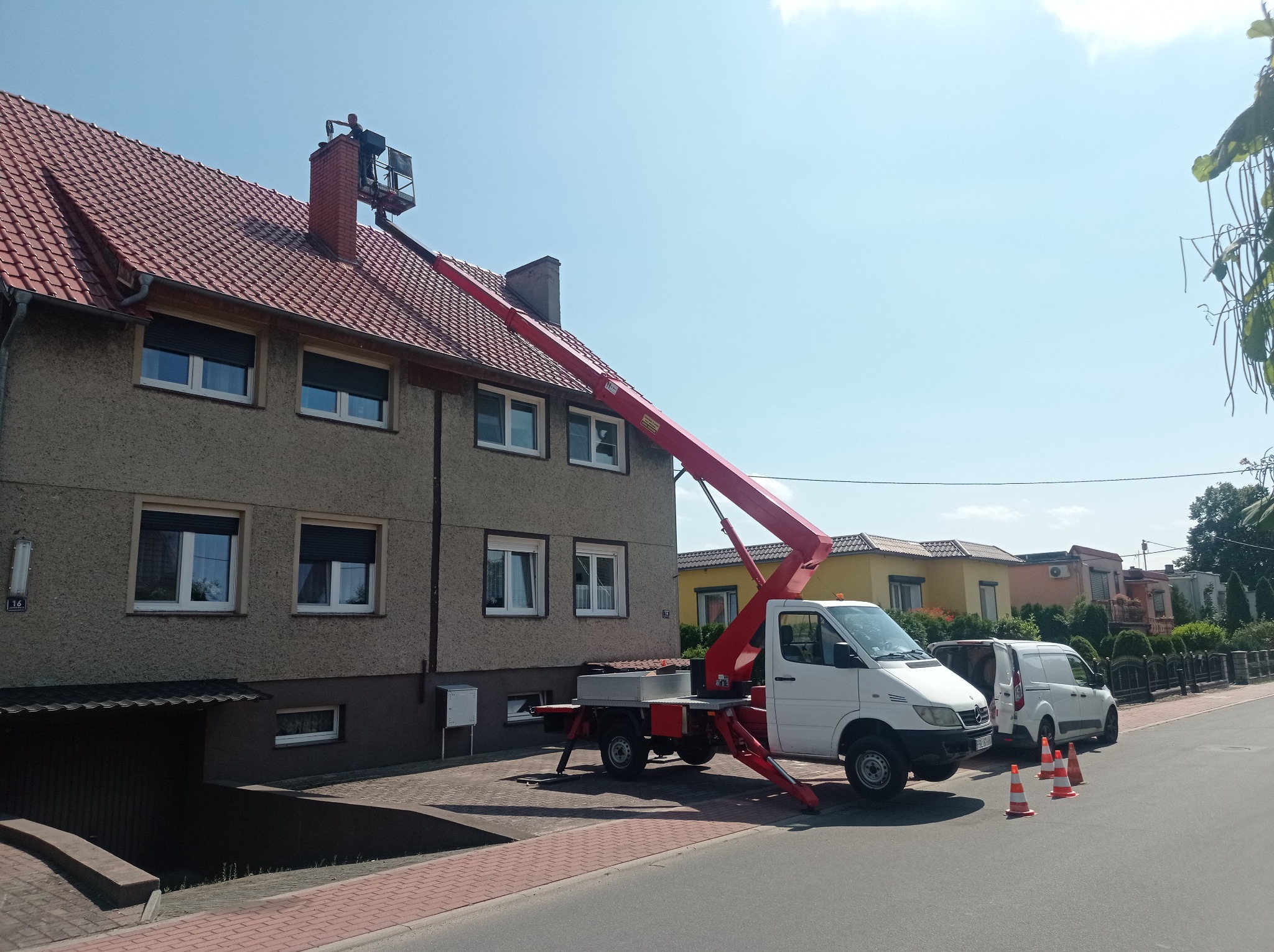 Pracownik na podnośniku koszowym przy kominie domu w Rzepinie, biały samochód dostawczy i pomarańczowe pachołki na ulicy.