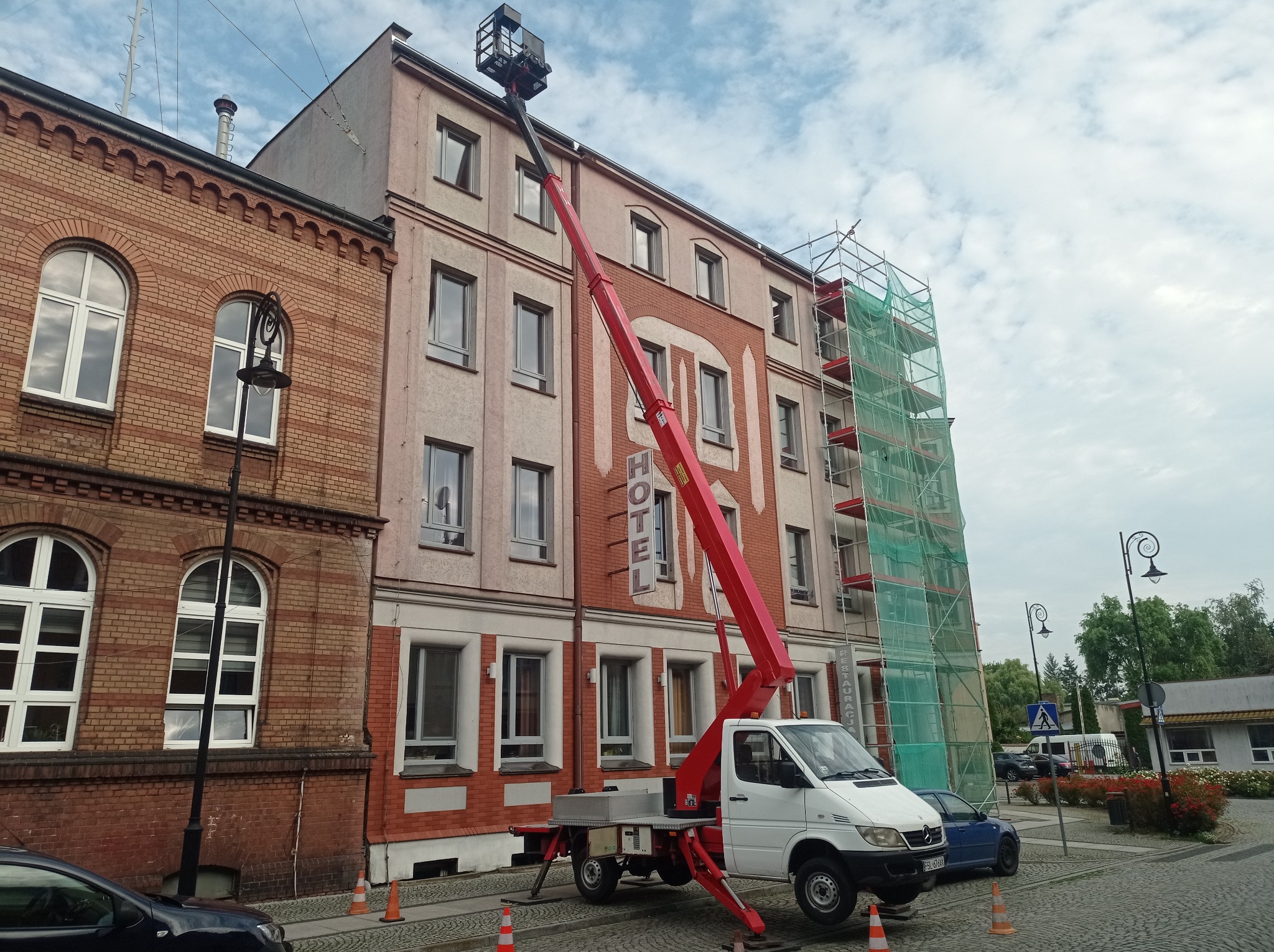 Czerwony podnośnik koszowy na białym samochodzie dostawczym ustawiony przed budynkiem hotelu z rusztowaniem, pomarańczowe pachołki na brukowanej ulicy.