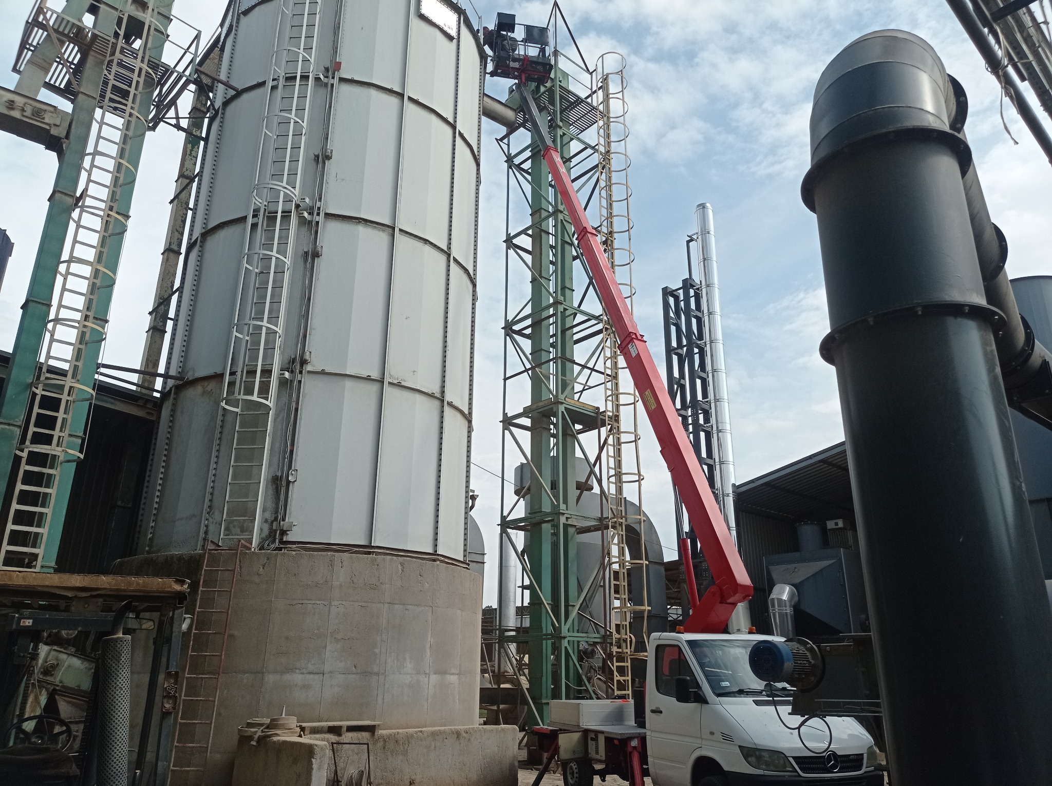 Praca dźwigowa przy montażu lub naprawie instalacji przemysłowej, widoczny biały samochód z czerwonym wysięgnikiem, silosy i elementy konstrukcyjne w tle.