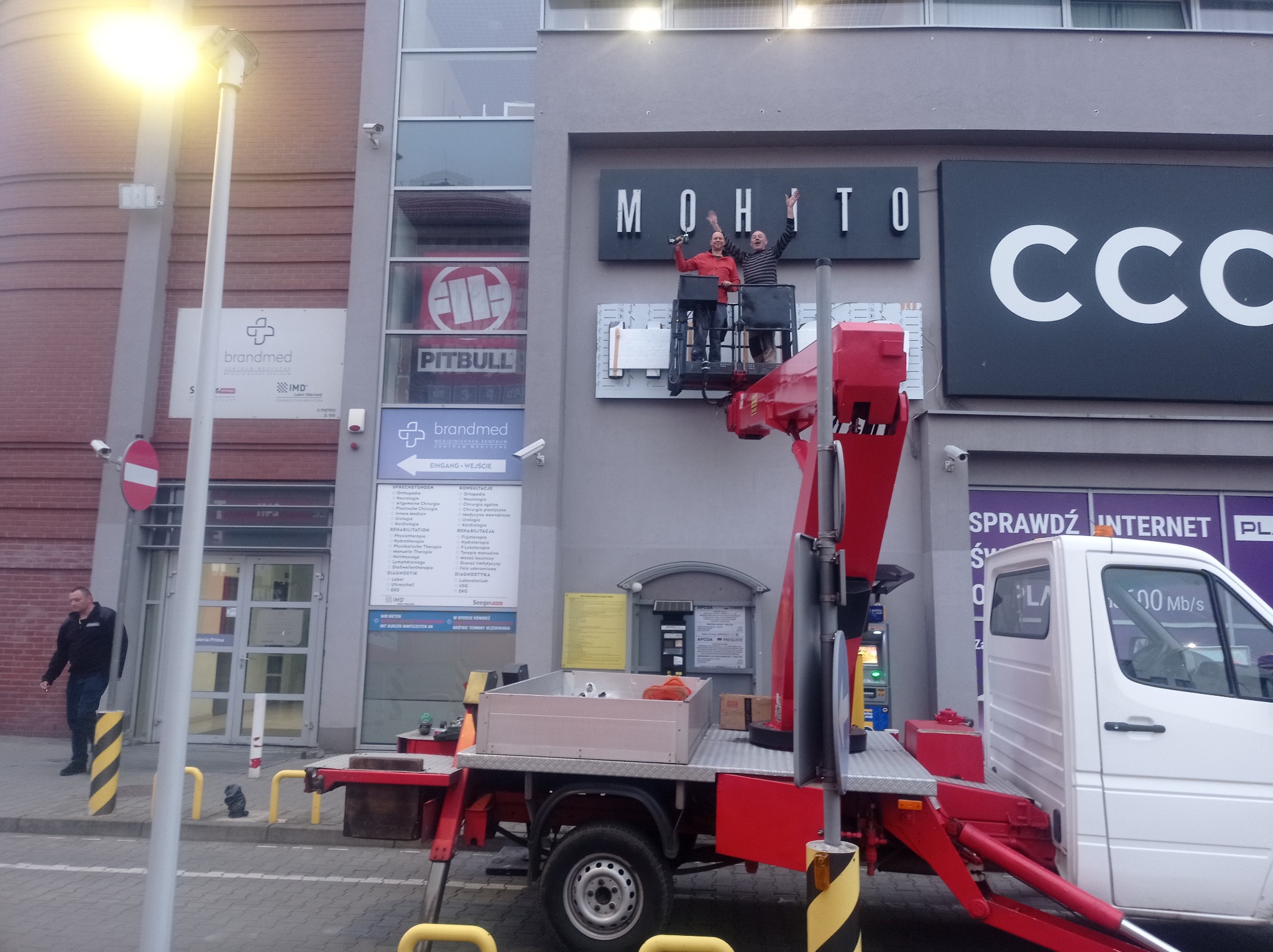 Dwóch mężczyzn w podnośniku koszowym montuje lub naprawia podświetlany szyld 'MOHITO' na elewacji budynku z innymi szyldami reklamowymi, widoczne narzędzia i elementy konstrukcyjne podnośnika.