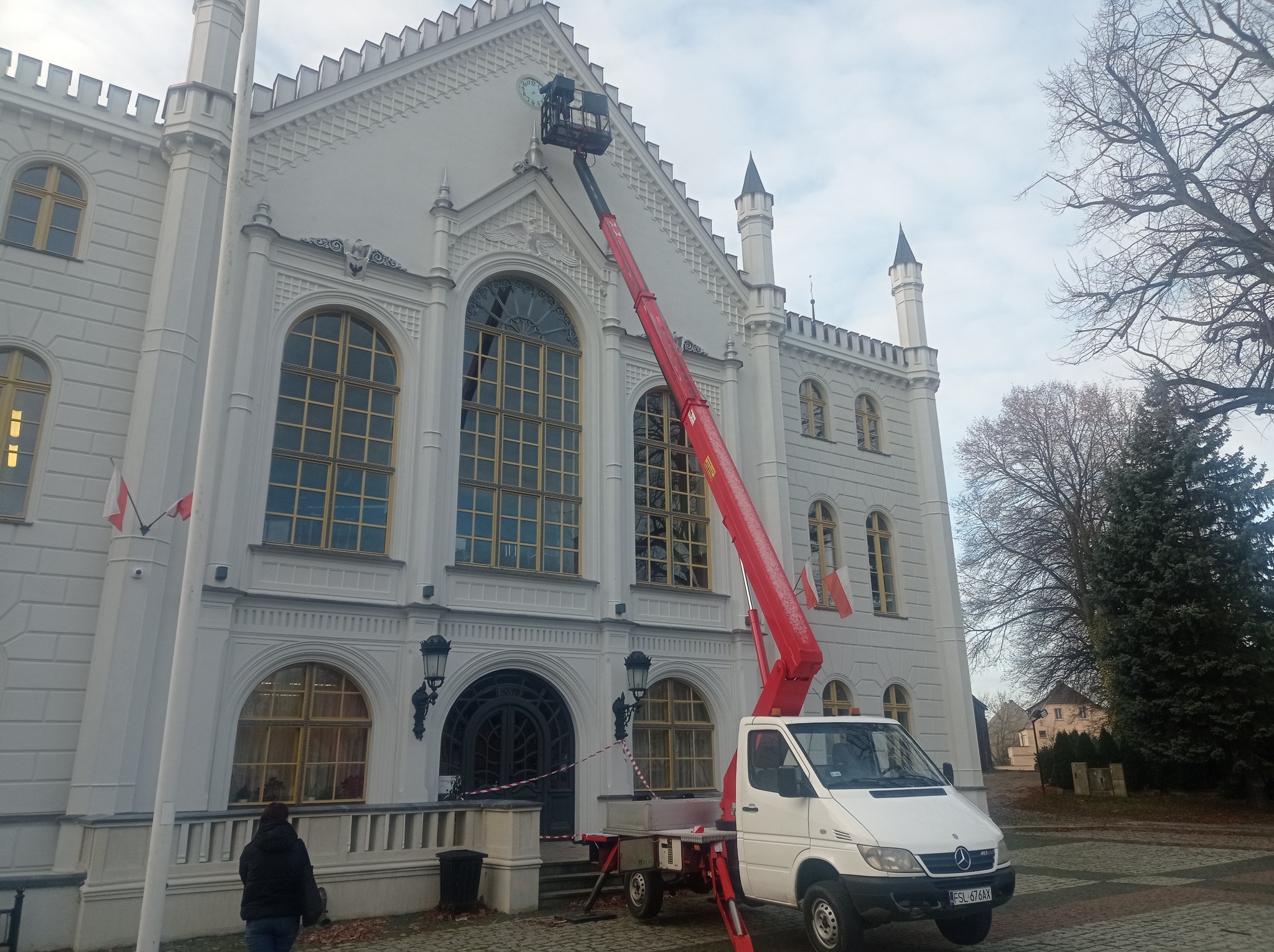 Prace na wysokościach przy zabytkowym, białym budynku z użyciem podnośnika koszowego; renowacja zegara na fasadzie.