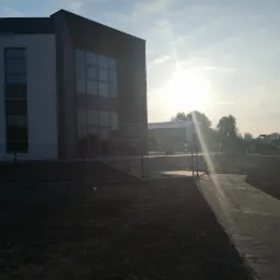 Nowoczesny budynek biurowy z przeszkloną fasadą i ogrodzeniem panelowym w trakcie montażu, widok na chodnik z kostki brukowej i okoliczne tereny w słoneczny dzień.