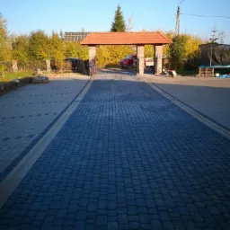 Szeroki podjazd z ciemnej kostki brukowej prowadzący przez kamienną bramę z czerwoną dachówką, w tle widoczne drzewa i fragment paneli słonecznych.