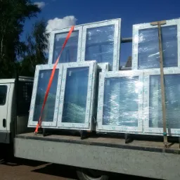 Transport okien PCV marki VEKA na pace białego samochodu dostawczego. Okna zabezpieczone folią i pomarańczowym pasem transportowym, błękitne niebo w tle.