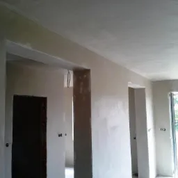 Wnętrze surowego domu po nałożeniu gładzi gipsowej na ściany i sufit, widoczne otwory drzwiowe i okienne, przygotowane do dalszych prac wykończeniowych.