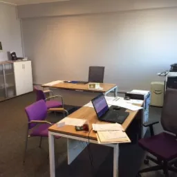 Wnętrze biura z dwoma biurkami, krzesłami w odcieniach fioletu, laptopem, dokumentami, drukarką i szafką w tle. Na biurku leżą dokumenty i telefon kom&oacute;rkowy. Oświetlenie dzienne.